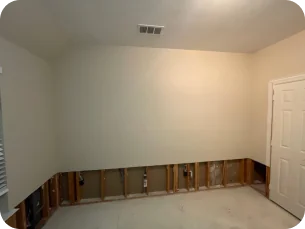 Interior room with beige walls and carpet removed, exposing wall studs and plumbing at the lower half.
