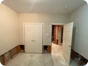 Empty room with beige walls, two doors, and lower wall panels removed exposing insulation and studs.