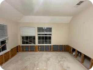 Empty room with beige walls, three windows with blinds, and lower wall panels removed exposing insulation.