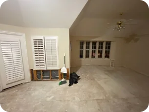 Empty room with bare concrete floor, partially removed lower wall panel near a door and window with white shutters, a broom, dustpan, and black trash bags on the floor.