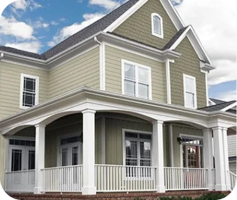 Large two-story green house with white trim, multiple windows, and a spacious wraparound porch under blue sky with clouds.