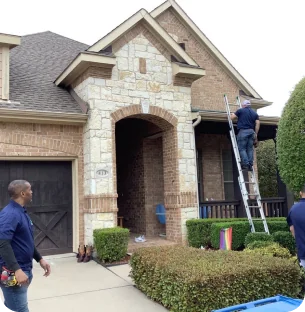 Two men working outside a brick and stone house, one on a ladder near the roof, the other standing on the driveway.