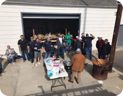Group of people standing and sitting outside a garage with wood and a table of cooking supplies, next to a large metal drum.