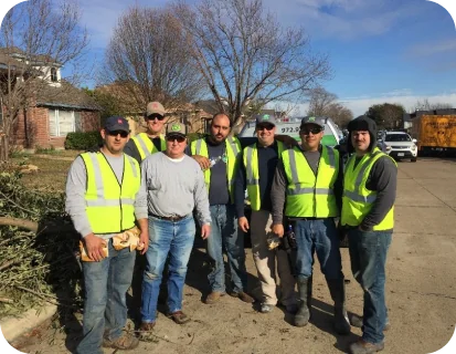 Seven men wearing yellow safety vests standing on a street in front of a house and vehicles on a sunny day.