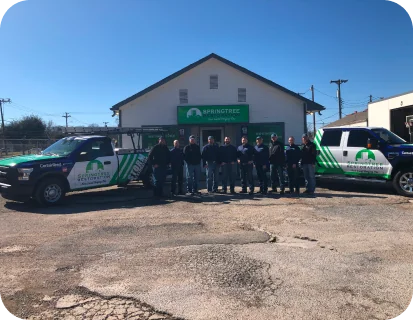 Group of twelve people standing between two branded service trucks in front of a Springtree lawn care building under a clear blue sky.