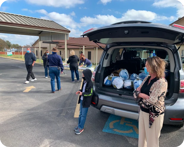 People wearing masks participate in a drive-through turkey distribution with turkeys loaded in the open trunk of an SUV parked in a handicapped spot.
