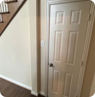 White interior door with four panels next to a light switch and staircase with wooden handrail and dark wood flooring.