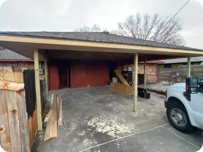 A partially covered driveway next to a house with scattered wooden planks and a white truck parked on the right side.