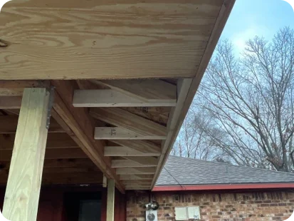 Close-up of wooden roof rafters under an overhang with a brick building and leafless trees in the background.