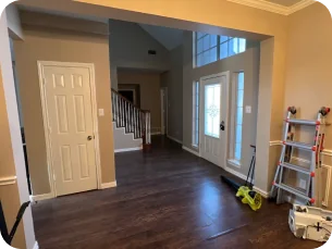 Empty room with dark wooden floor, white door, staircase, large front door with sidelights, and cleaning tools against the wall.