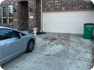 Concrete driveway with scattered dry leaves in front of a brick house with a white garage door and a green trash bin, next to a parked silver car.