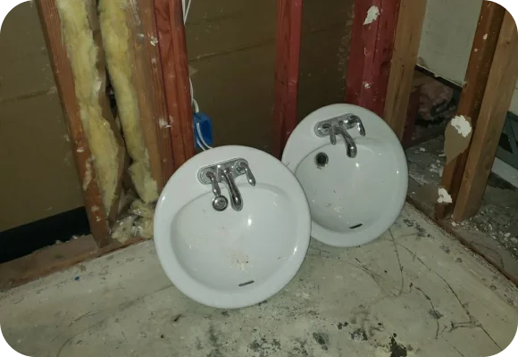 Two white wall-mounted sinks placed on a dirty floor in a partially constructed or demolished room with exposed wooden studs and insulation.