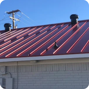 Red metal roof with two black vents and part of a brick building under a clear blue sky.