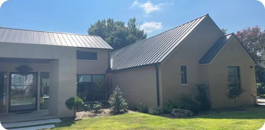 Single-story modern house with cream-colored brick walls, metal roofs, and a landscaped front yard under a clear blue sky.