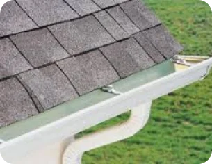 Close-up of a roof edge with gray asphalt shingles and a white gutter system against a grassy background.