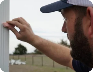 Man wearing a cap touching or cleaning a glass window with an outdoor rural landscape in the background.