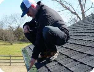 Man wearing a cap and dark clothing crouching on a shingled roof inspecting the gutter.