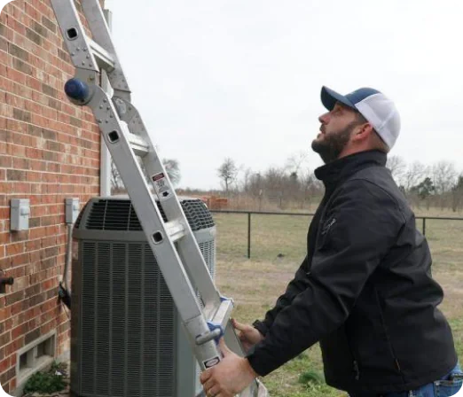 Man in a black jacket and baseball cap setting up a metal ladder against a brick building near an air conditioning unit.
