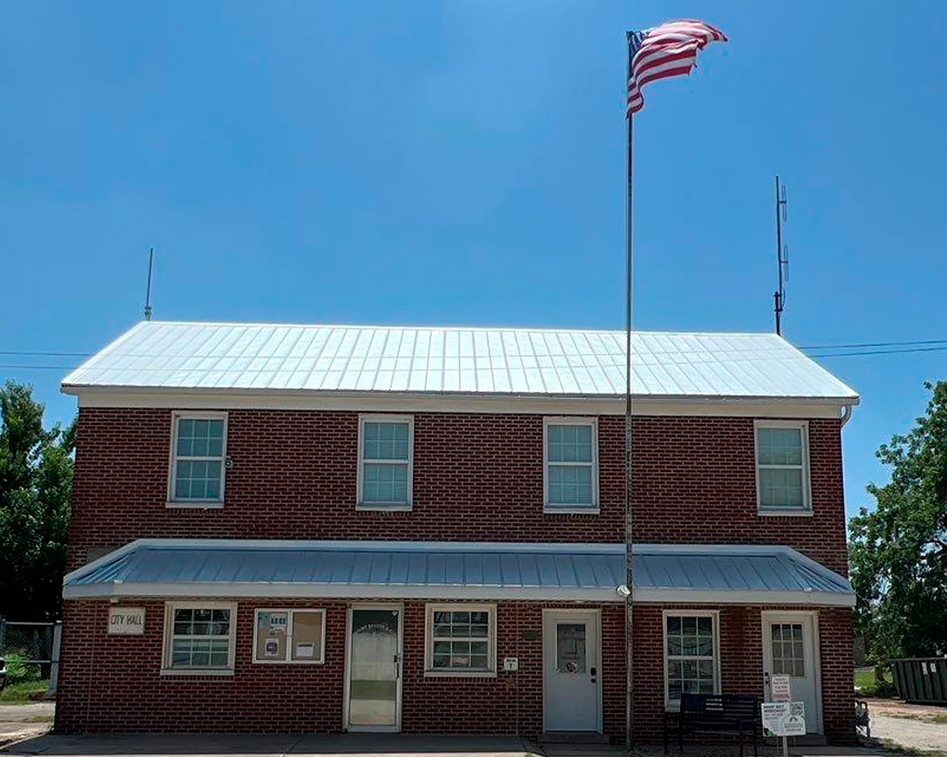 Two-story brick city hall building with a white metal roof and an American flag on a tall flagpole.