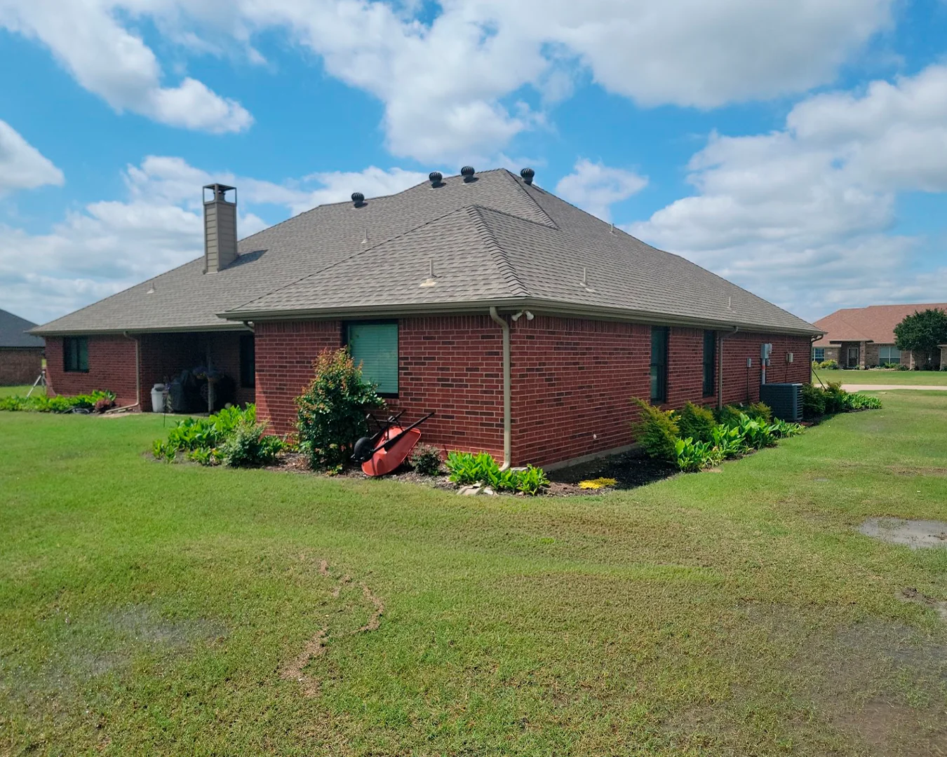 Brick house with a large sloped roof, surrounding green lawn, and garden beds with plants along the sides under a blue sky with scattered clouds.