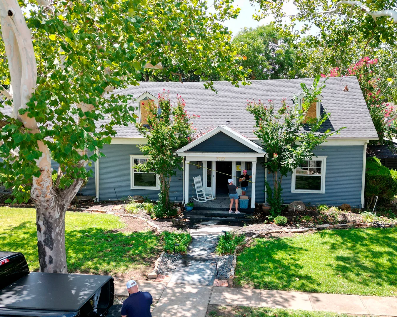 Blue house with white trim and a gray roof, two people standing on the front porch holding a measuring tape, and a man working near a black pickup truck under a tree.