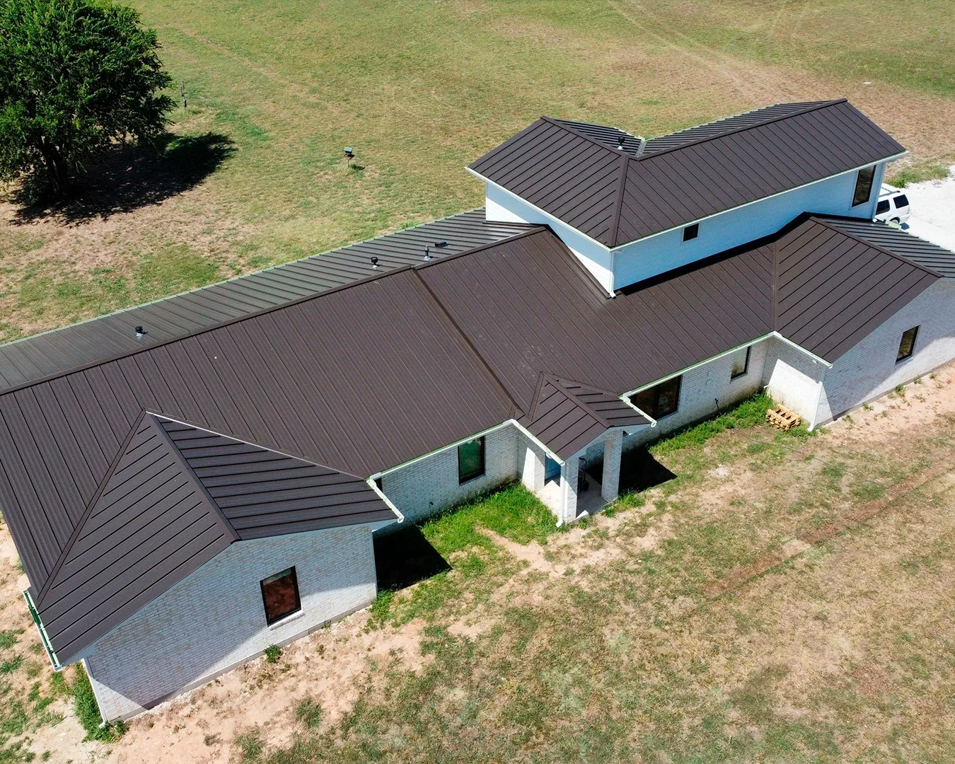 Aerial view of a large single-story white brick house with a dark brown metal roof and surrounding dry grass.