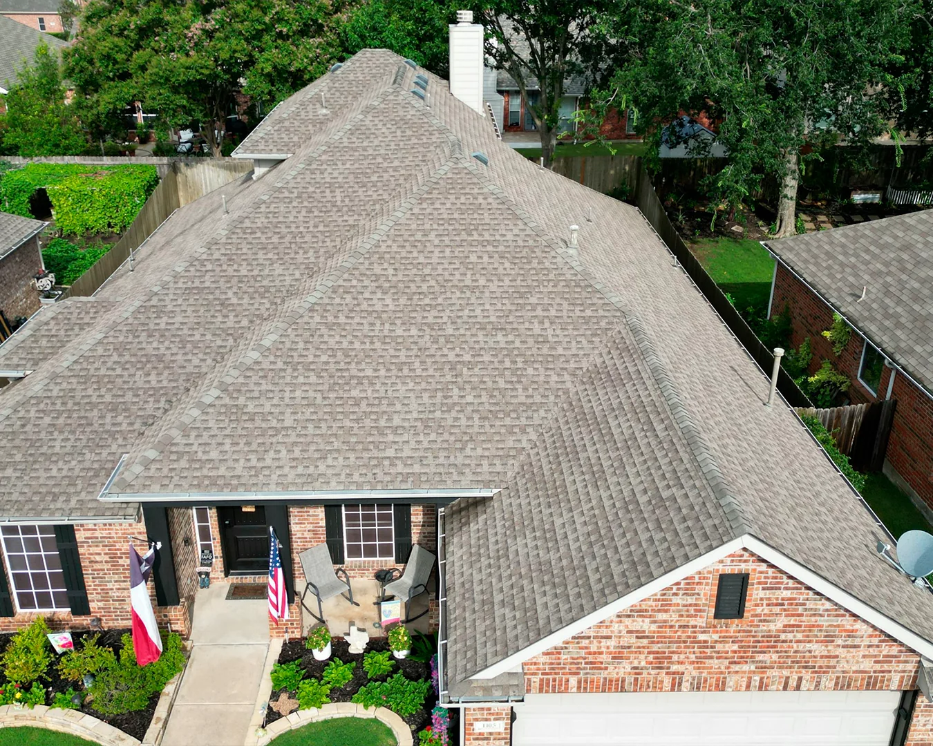 Aerial view of a brick suburban house with a large gray shingled roof, front porch with chairs, and American and Texas flags.