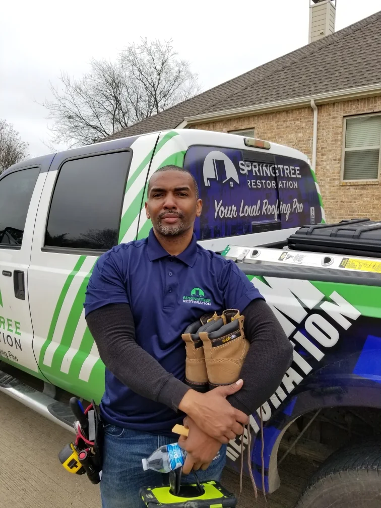 Man in a blue SpringTree Restoration polo shirt standing in front of a SpringTree Restoration work truck holding tan work boots, a water bottle, and a tool case.