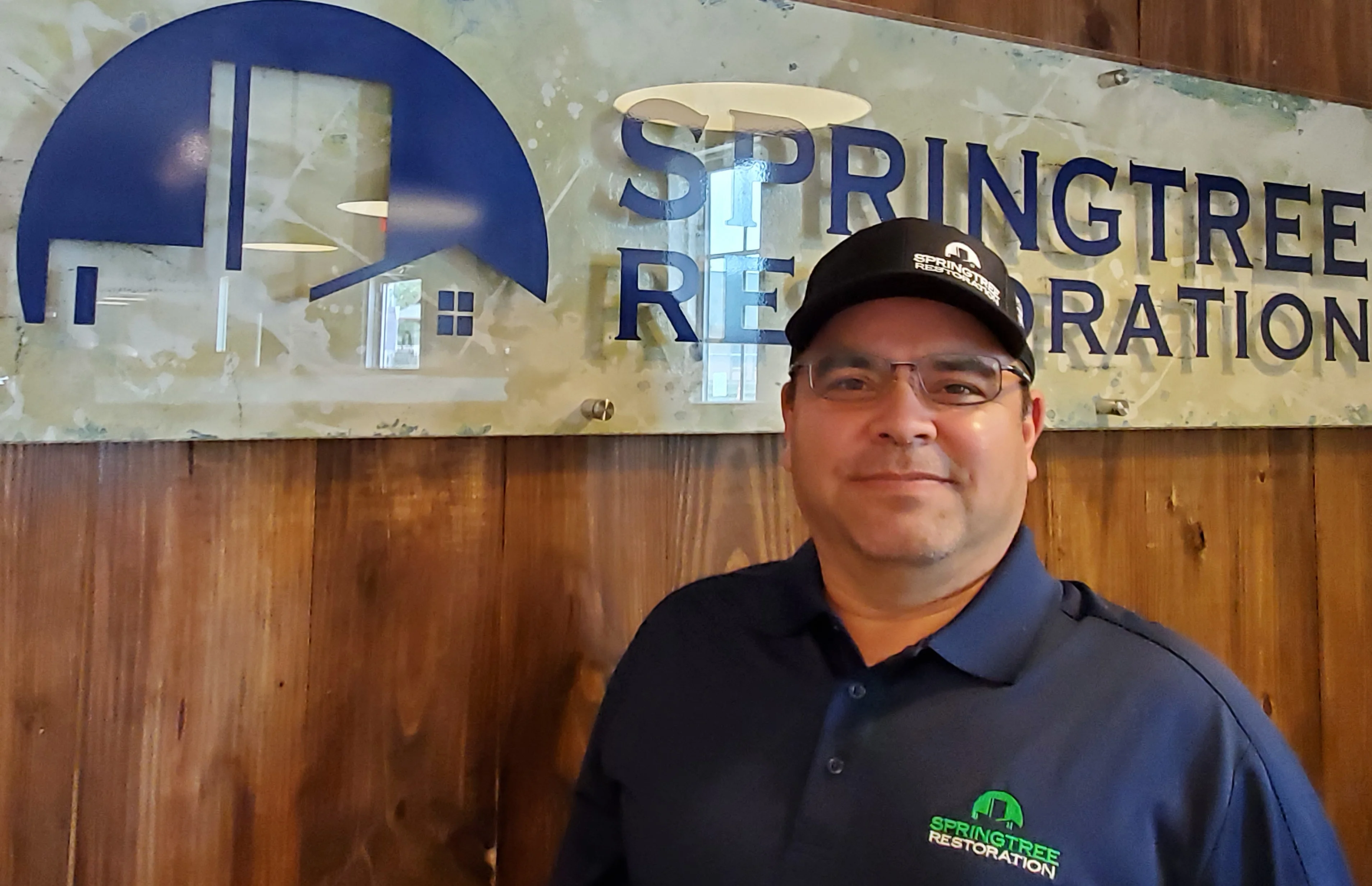 Man wearing glasses, a black Springtree Restoration cap, and a navy blue polo shirt with the Springtree Restoration logo, standing in front of a wooden wall with a large Springtree Restoration sign.