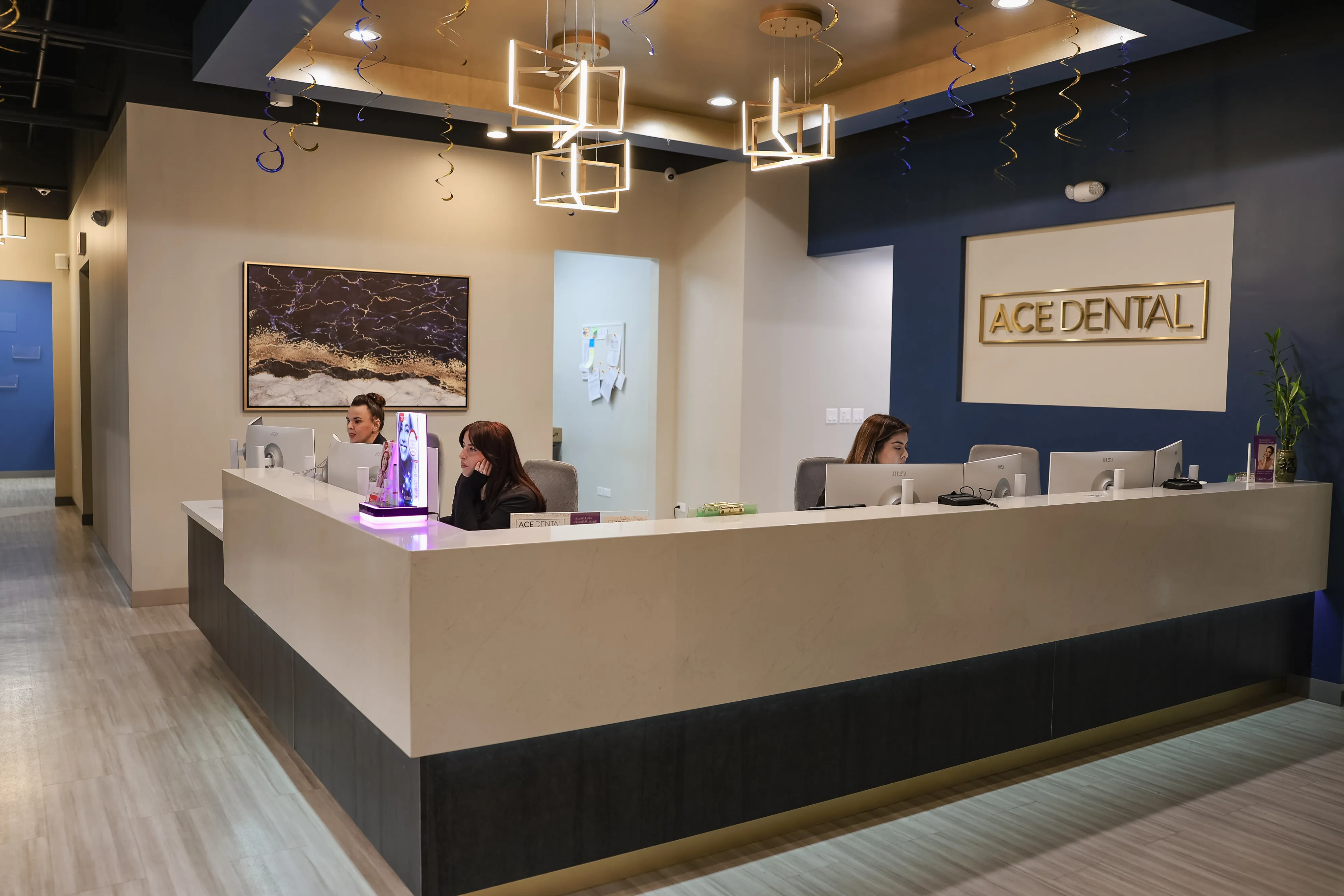 Reception area at Ace Dental with three staff members working behind a modern white and dark wood desk under geometric pendant lights.