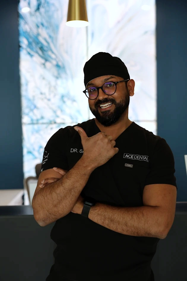 Smiling male dentist wearing glasses, black scrubs, and a black cap, giving a thumbs-up gesture inside a dental office.