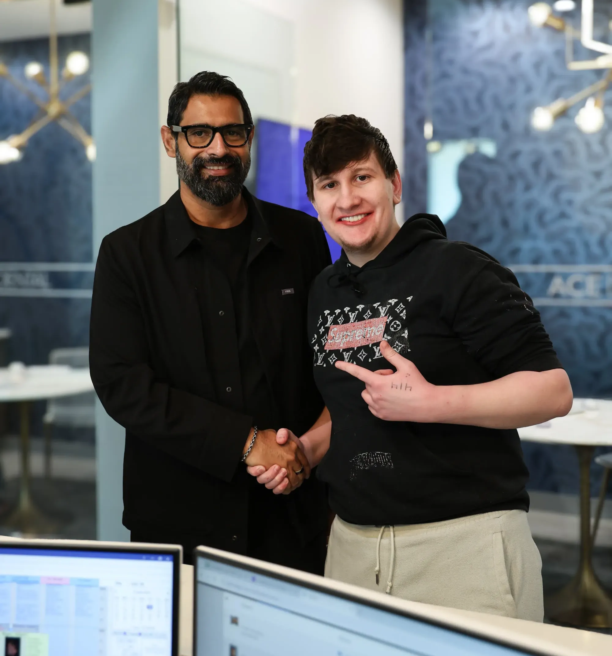 Two men smiling and shaking hands in an office setting, one wearing glasses and black jacket, the other in a black Supreme hoodie.