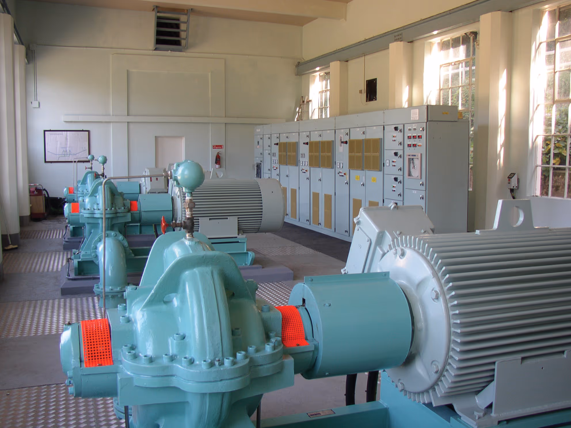 Interior of an industrial facility showing a row of large blue pumps connected to electric motors with control panels along the wall.