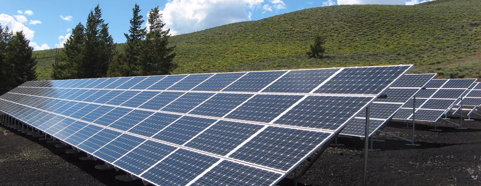 Rows of solar panels installed on the ground with green hills and trees in the background under a partly cloudy sky.