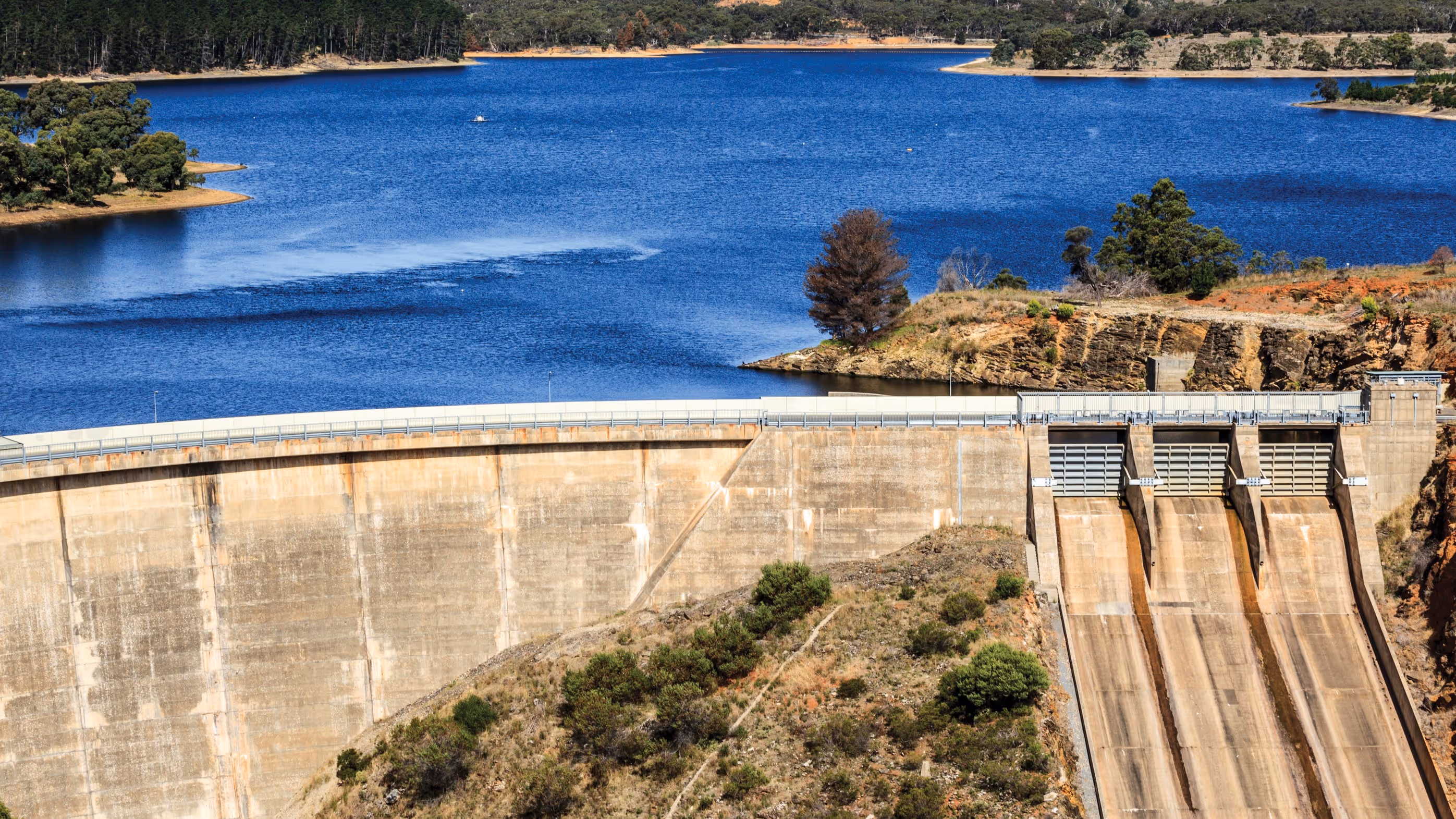Concrete dam with a reservoir of blue water and surrounding trees and hills.