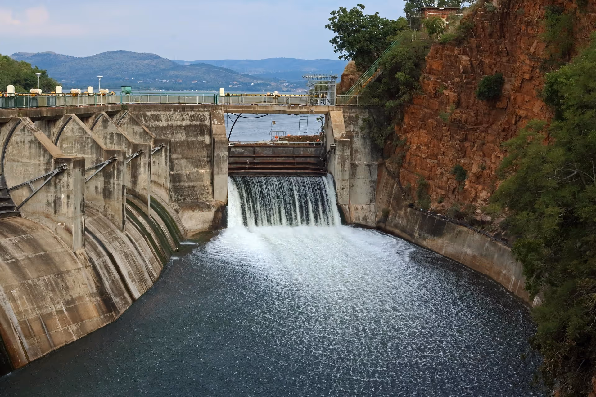 Concrete dam releasing water into a river with rocky cliffs and trees on one side and mountains in the background.