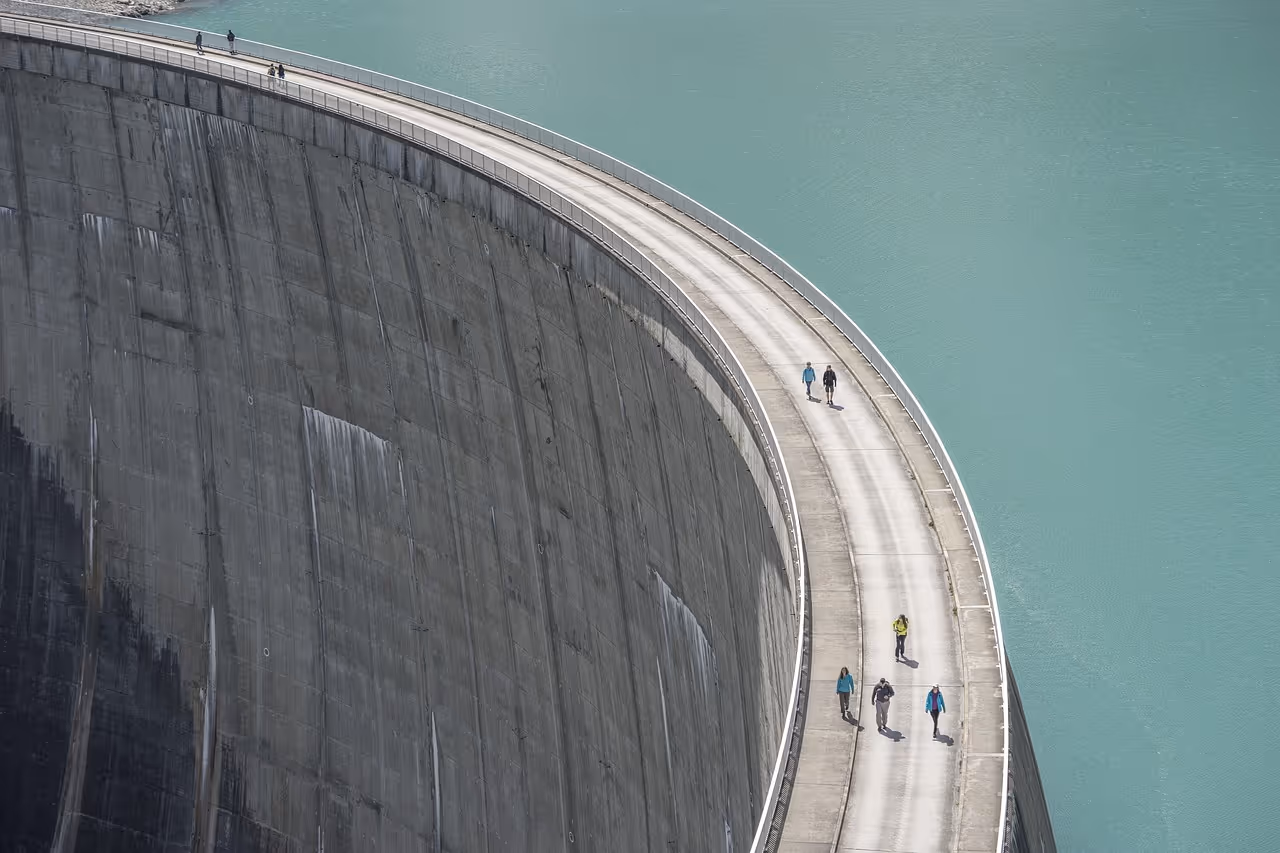 People walking on top of a large curved concrete dam with turquoise water on one side.