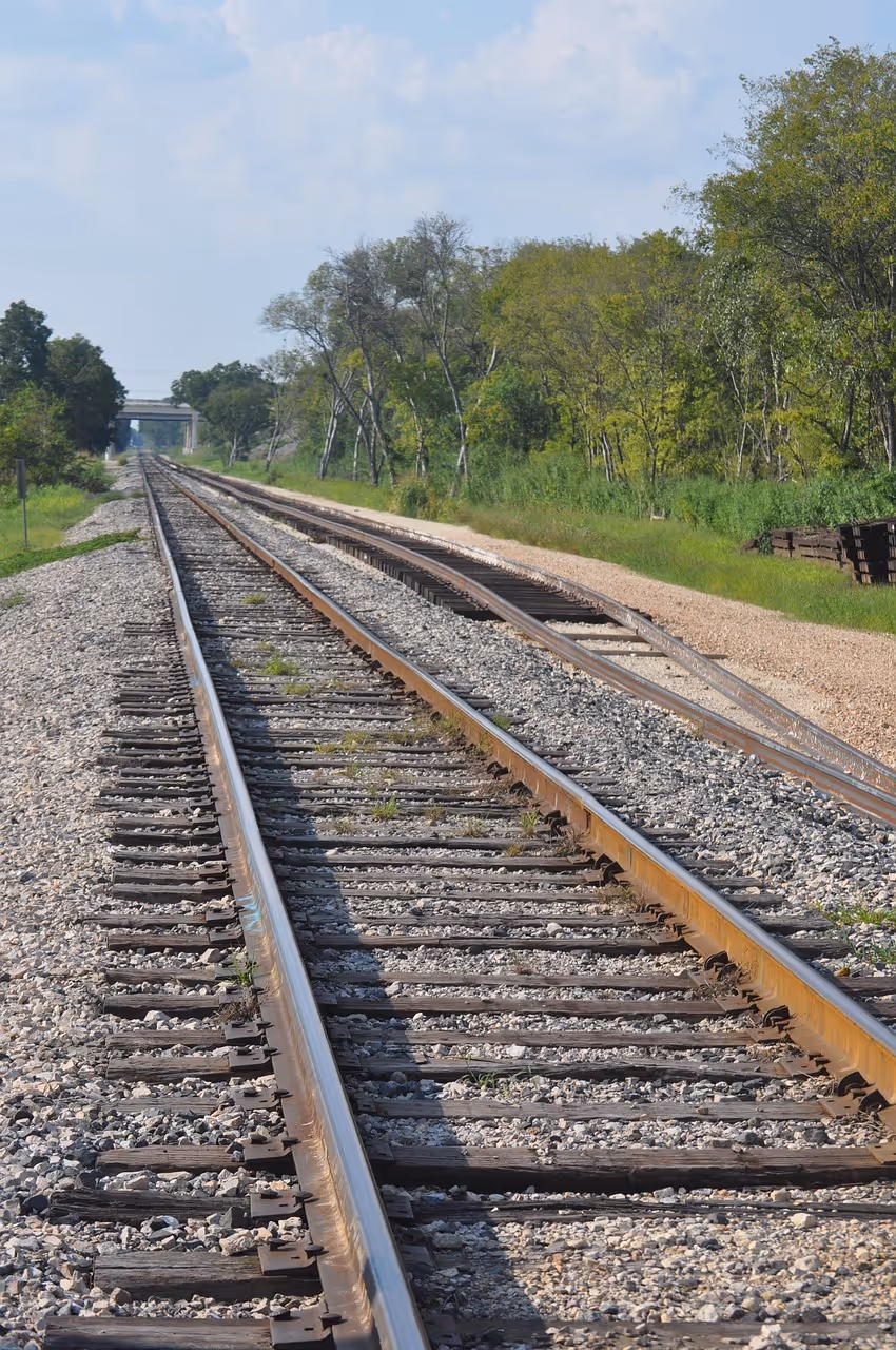 Two railroad tracks running through a rural area with trees and a bridge in the distance under a partly cloudy sky.