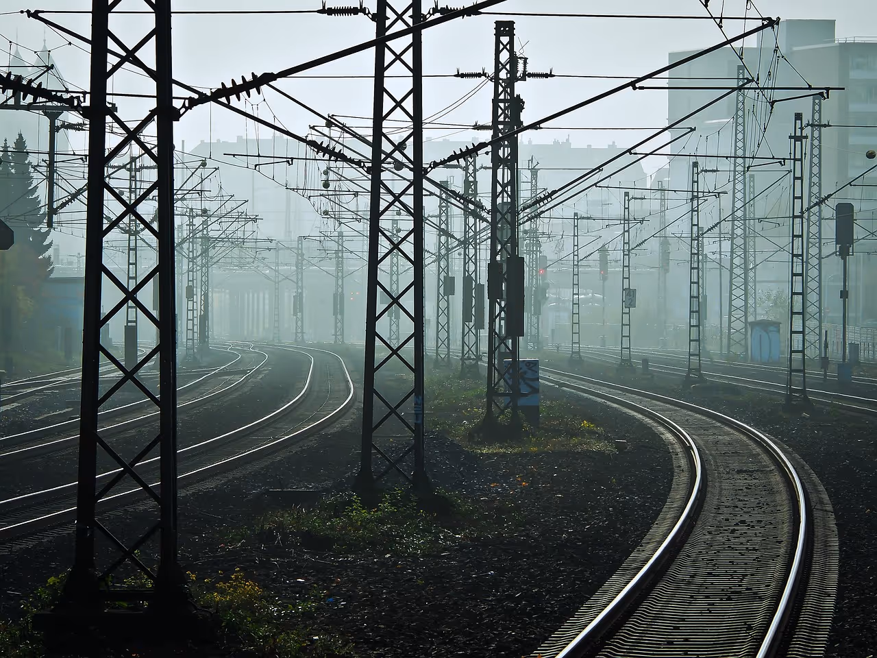 Foggy railway tracks with overhead electric wires and support poles stretching into the distance near urban buildings.