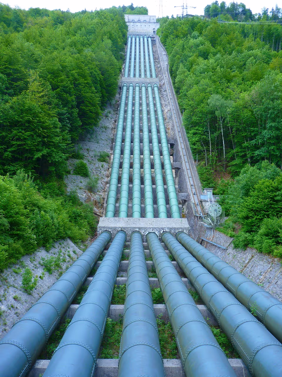 Large green pipeline system running down a steep forested hillside.