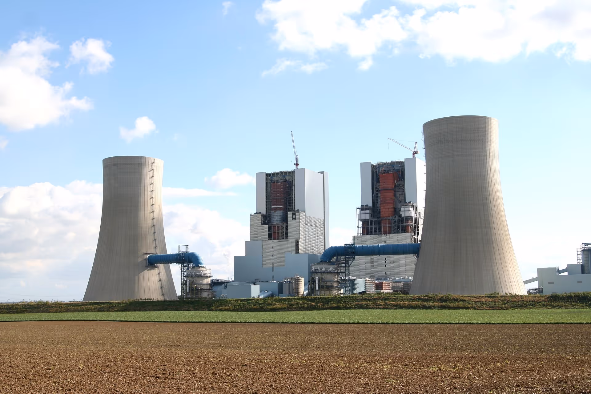 Power plant with two large cooling towers connected by blue pipes under a partly cloudy sky.