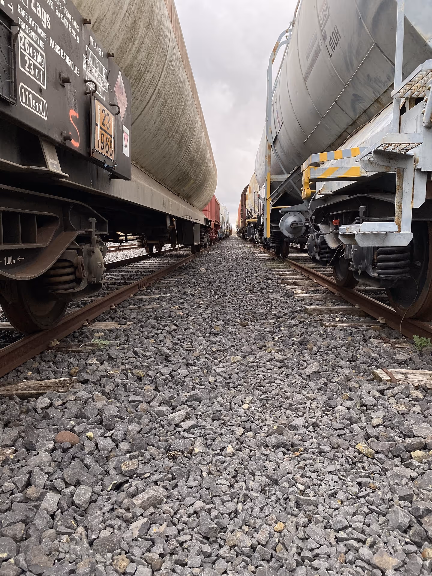 Close-up view of gravel between railway tracks with freight train tank cars on both sides under a cloudy sky.