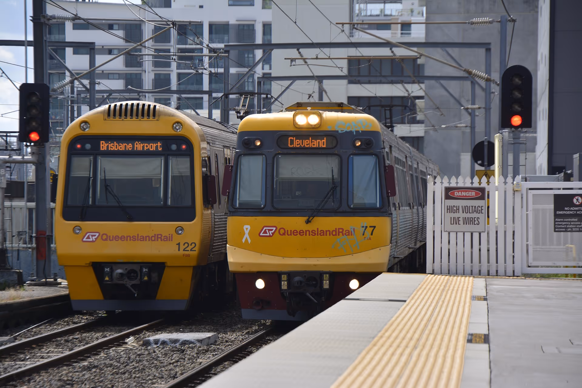 Two yellow QueenslandRail trains stopped side by side at a station, one headed to Brisbane Airport and the other to Cleveland.