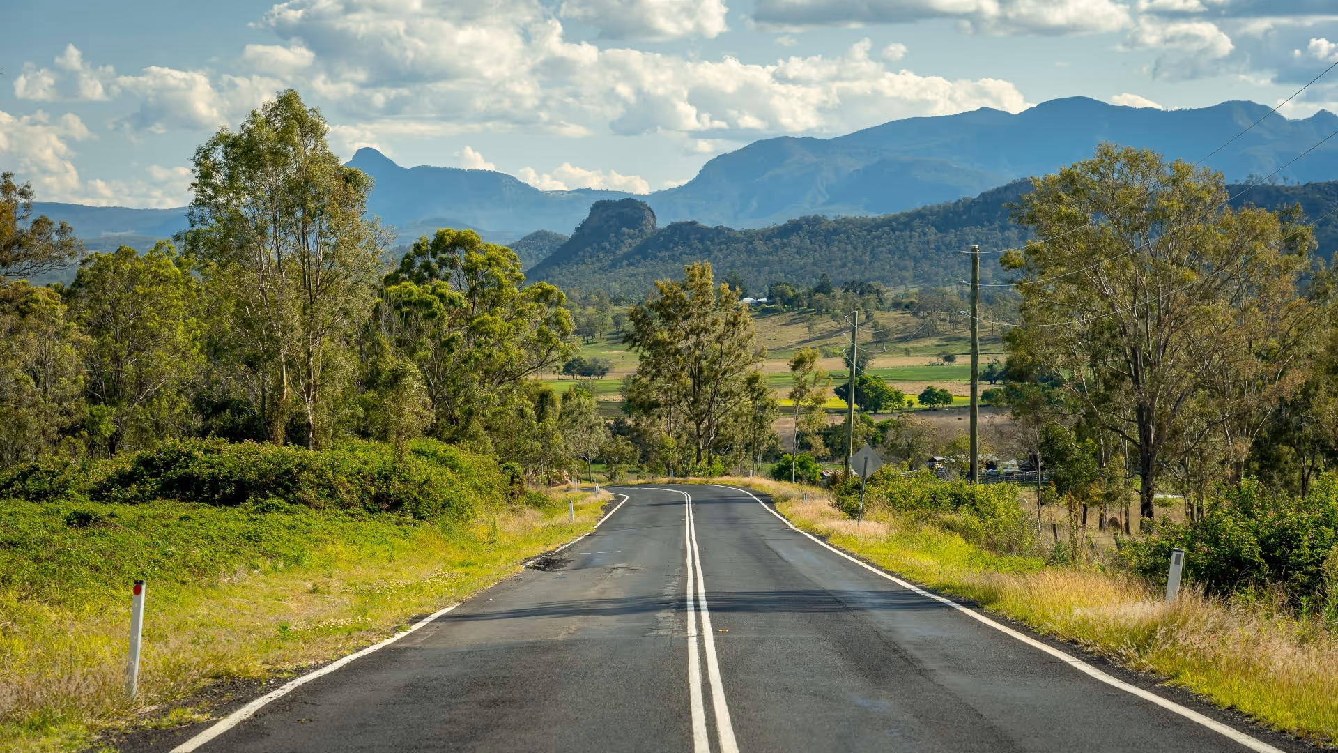 Two-lane rural road with double white lines stretching into a scenic landscape of green trees and mountains under a partly cloudy sky.