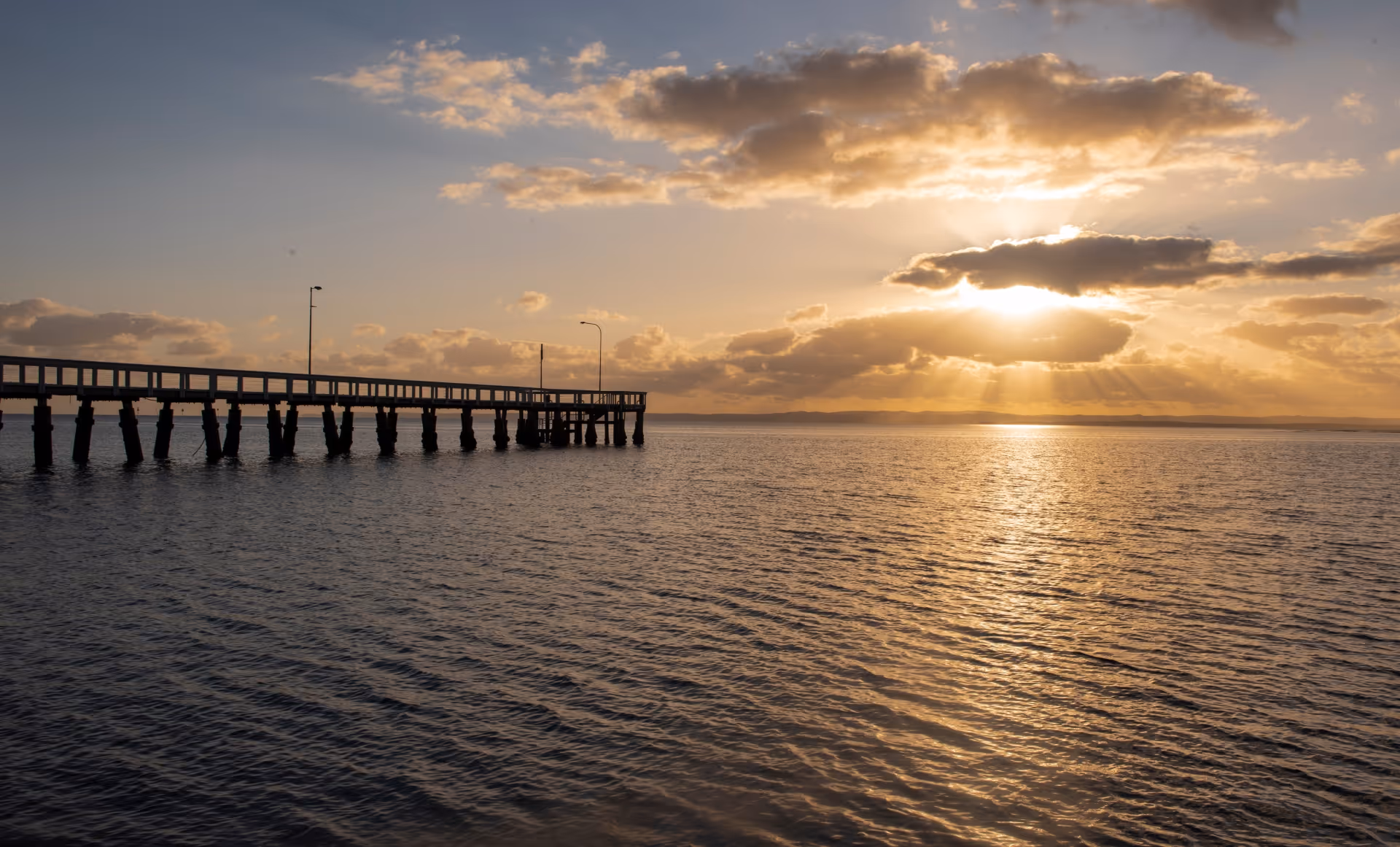 Wooden pier extending into calm water at sunset with sun rays breaking through clouds.