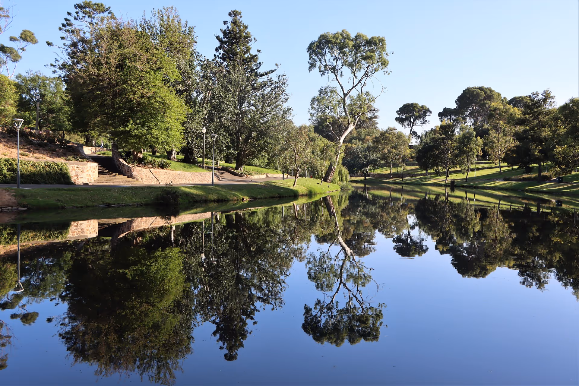 Calm river reflecting lush green trees and a clear blue sky in a park setting.