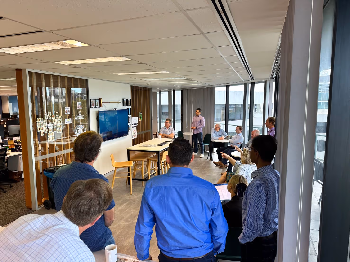 Office meeting with a group of people standing and sitting around a screen displaying a presentation.