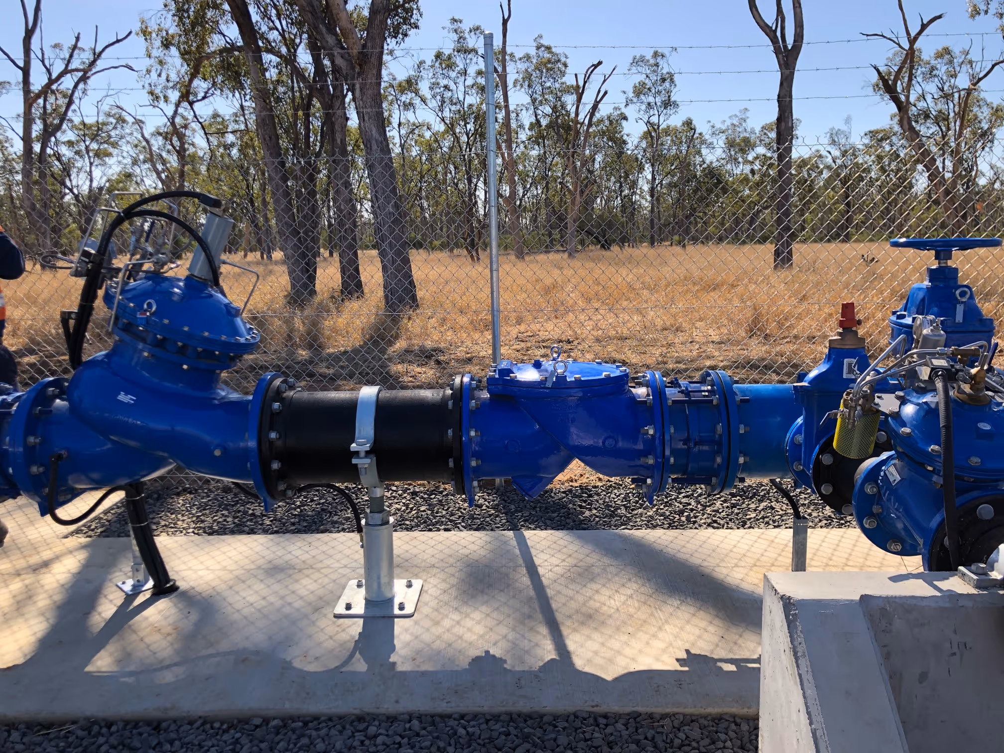 Blue industrial water pipe assembly with valves on a concrete platform surrounded by a chain-link fence and trees in the background.