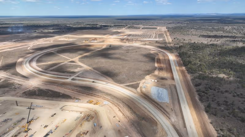 Aerial view of a large construction site with partially built curved roads and cleared land extending to a forested area under a blue sky.