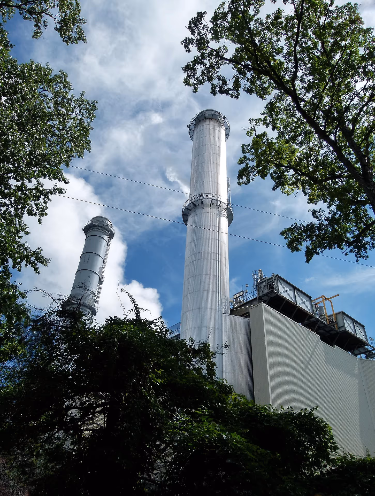 Tall industrial smokestacks surrounded by green trees under a partly cloudy blue sky.